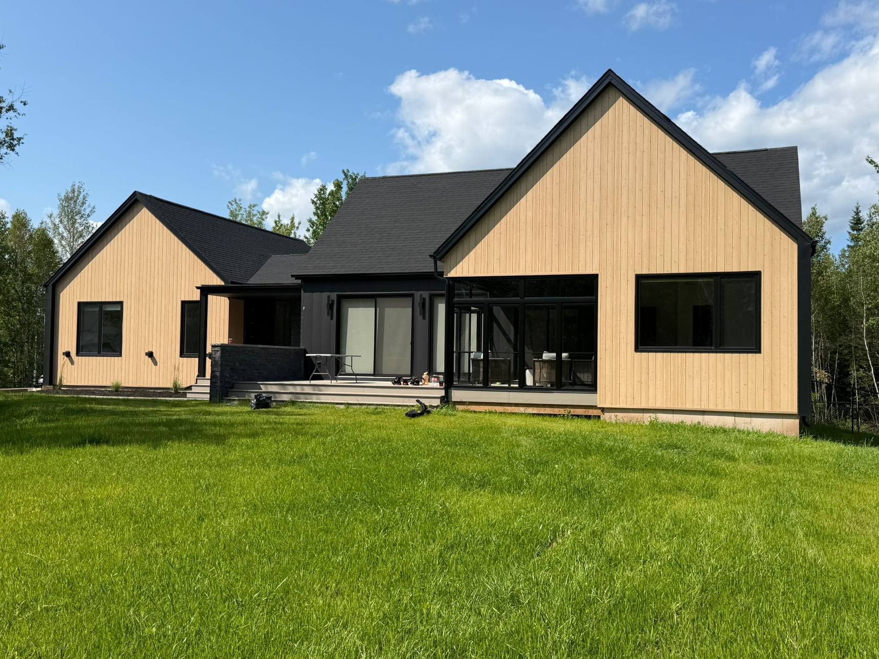 A completed custom home with vertical cedar-toned siding, black trim and a black gable roof, seen from the back lawn